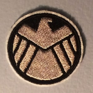 May include: A round embroidered patch with a white border and a black background. The patch features a gold eagle with a shield design.