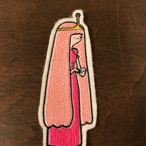 May include: Embroidered iron-on patch featuring a pink-haired cartoon princess wearing a pink dress and a gold crown.