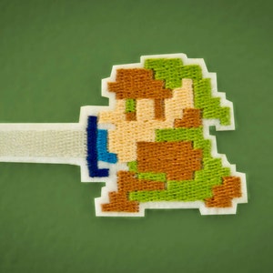 May include: A green, brown, and blue embroidered patch of a video game character holding a sword. The character is wearing a green tunic and brown pants.