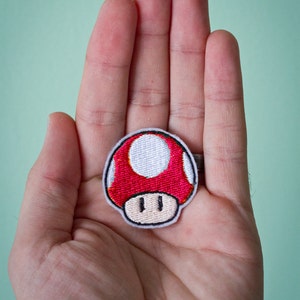 May include: A red and white embroidered mushroom patch with a white circle in the center. The patch is held in a person's hand.