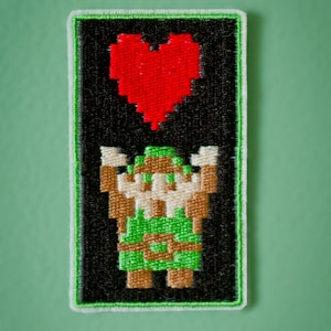 May include: A green pixelated character holding a red pixelated heart. The character and heart are embroidered on a black background with a white border.
