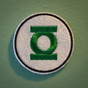 May include: A white and green embroidered patch with the Green Lantern logo. The logo is a stylized letter "O" with a green ring around it and a green bar above and below the ring.