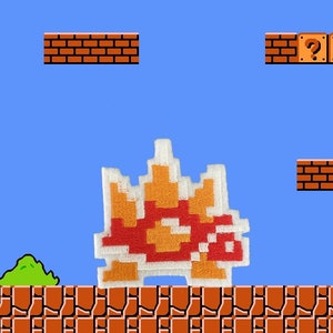May include: Embroidered patch featuring a pixelated, retro video game character in shades of red, orange, and white. The patch is set against a blue background with brick platforms and question mark blocks, reminiscent of a classic platformer game.