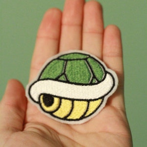 Puede incluir: Un parche bordado de un caparazón de tortuga, con colores verde y amarillo y un borde blanco. El parche está en la mano de una persona.