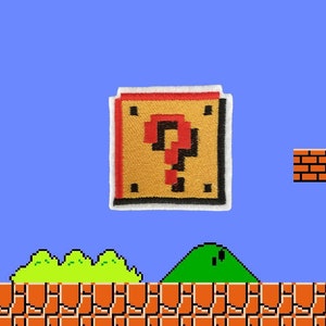 May include: A pixelated embroidered patch of a yellow question mark block from the Super Mario Bros. video game. The block has a red question mark and a black outline. The patch is on a blue background.