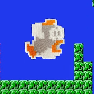 May include: A white and orange embroidered patch of a video game character, a grey and white creature with orange feet and a small orange hat, against a blue background.