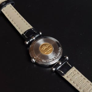 May include: A wristwatch with a black leather strap featuring a beige underside and a silver-tone metal case back. The case back is engraved with the text Van Cleef & Arpels Paris and La collection, and features a central gold-tone circular emblem.