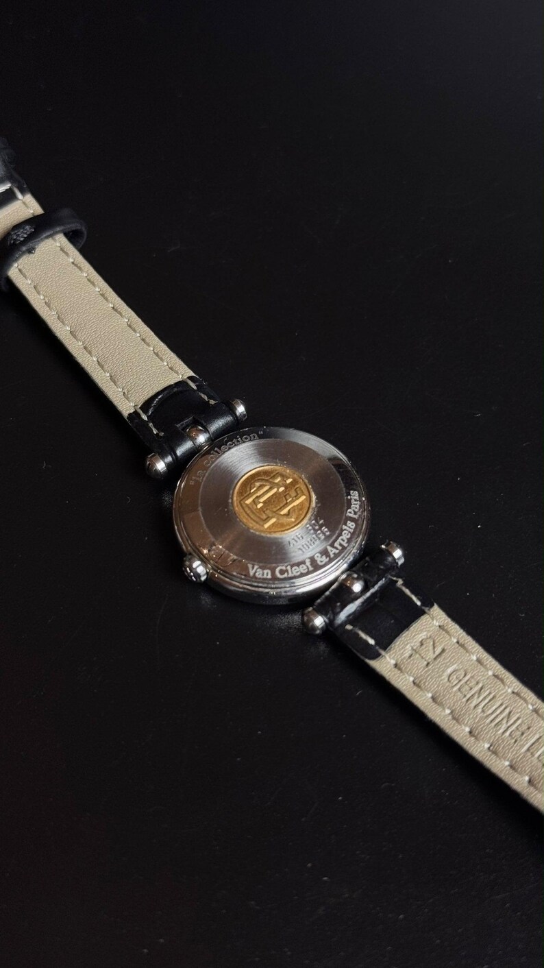 May include: A wristwatch with a black leather strap featuring a beige underside and a silver-tone metal case back. The case back is engraved with the text Van Cleef & Arpels Paris and La collection, and features a central gold-tone circular emblem.