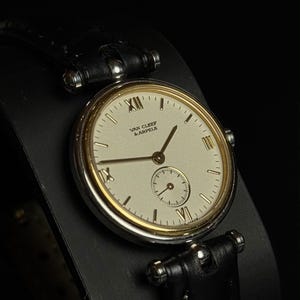 May include: A wristwatch with a round silver-tone case and a gold-tone bezel. The cream-colored dial features the text "VAN CLEEF & ARPELS," gold-tone hands, Roman numerals at the 12, 6, and 9 o'clock positions, and a small sub-dial. It has a black leather strap.