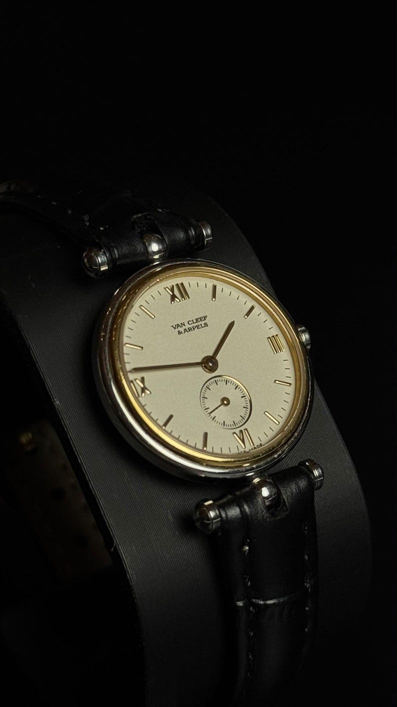 May include: A wristwatch with a round silver-tone case and a gold-tone bezel. The cream-colored dial features the text "VAN CLEEF & ARPELS," gold-tone hands, Roman numerals at the 12, 6, and 9 o'clock positions, and a small sub-dial. It has a black leather strap.