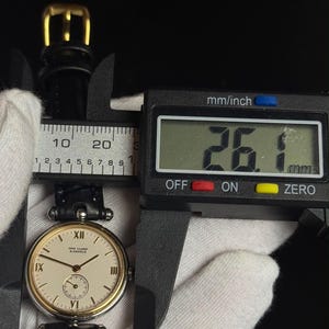 May include: A Van Cleef & Arpels wristwatch with a round cream-colored dial, gold-tone bezel, and black leather strap. A digital caliper measures the watch case at 26.1 mm. The dial features black Roman numerals at the 12, 9, 6, and 3 positions.