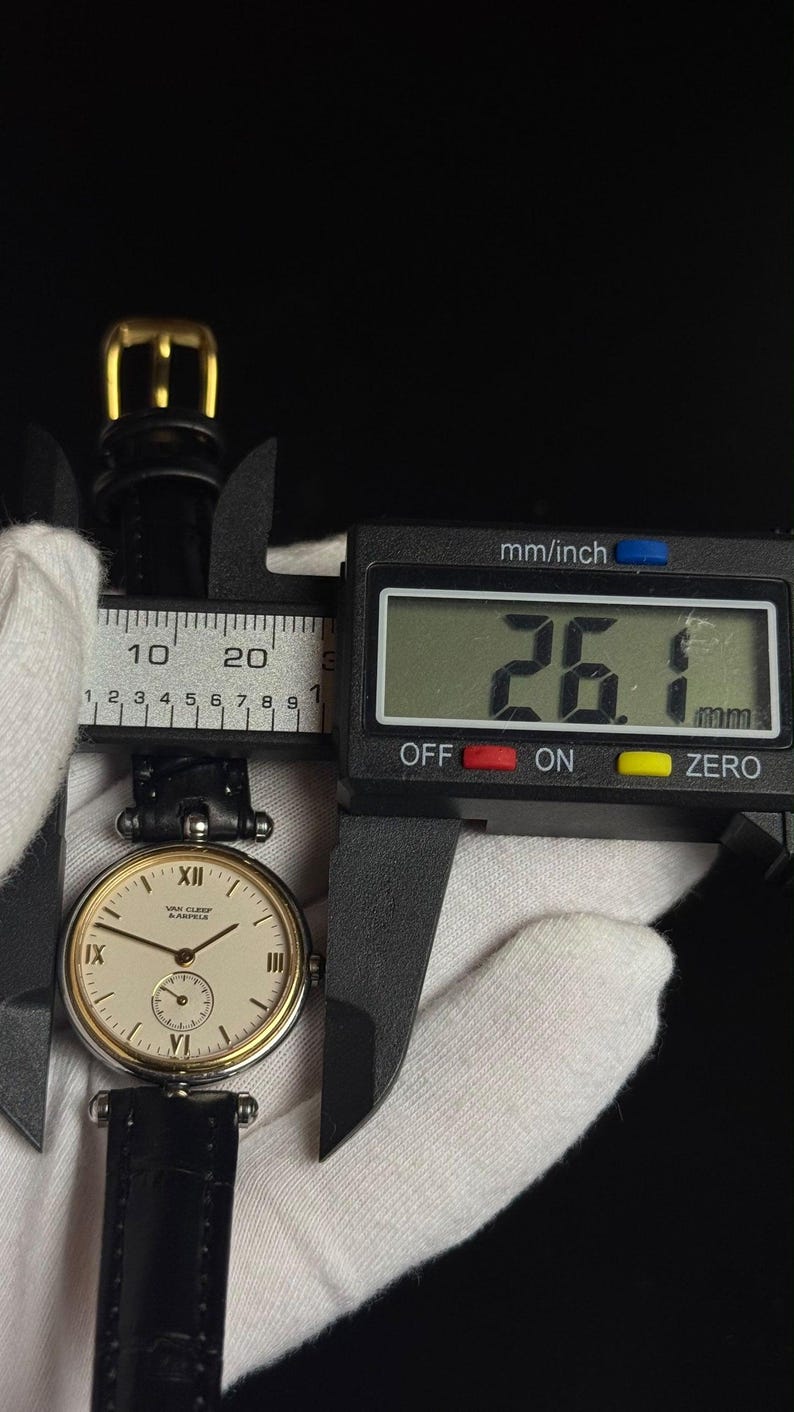 May include: A Van Cleef & Arpels wristwatch with a round cream-colored dial, gold-tone bezel, and black leather strap. A digital caliper measures the watch case at 26.1 mm. The dial features black Roman numerals at the 12, 9, 6, and 3 positions.