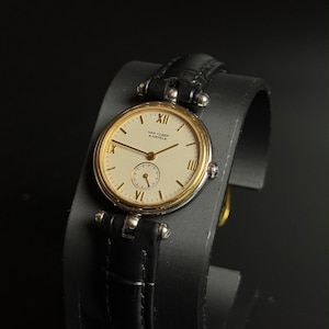 May include: A wristwatch with a round gold-tone bezel and a cream-colored dial featuring the text Van Cleef & Arpels. The dial includes black Roman numeral hour markers at the twelve, three, six, and nine positions, thin hands, and a sub-dial.