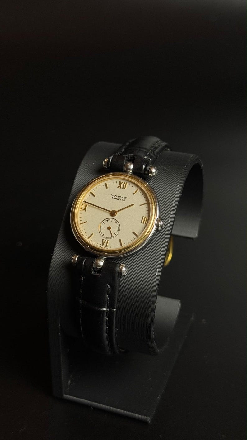 May include: A wristwatch with a round gold-tone bezel and a cream-colored dial featuring the text Van Cleef & Arpels. The dial includes black Roman numeral hour markers at the twelve, three, six, and nine positions, thin hands, and a sub-dial.