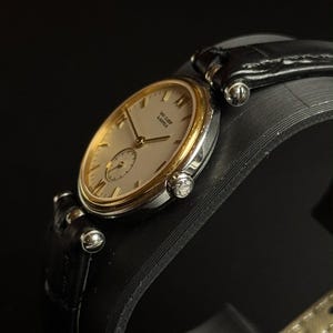 May include: A wristwatch featuring a round silver-tone case with a gold-tone bezel, a cream-colored dial with gold-tone hands and indices, and the text Van Cleef & Arpels. The watch includes a black leather strap with silver-tone hardware.