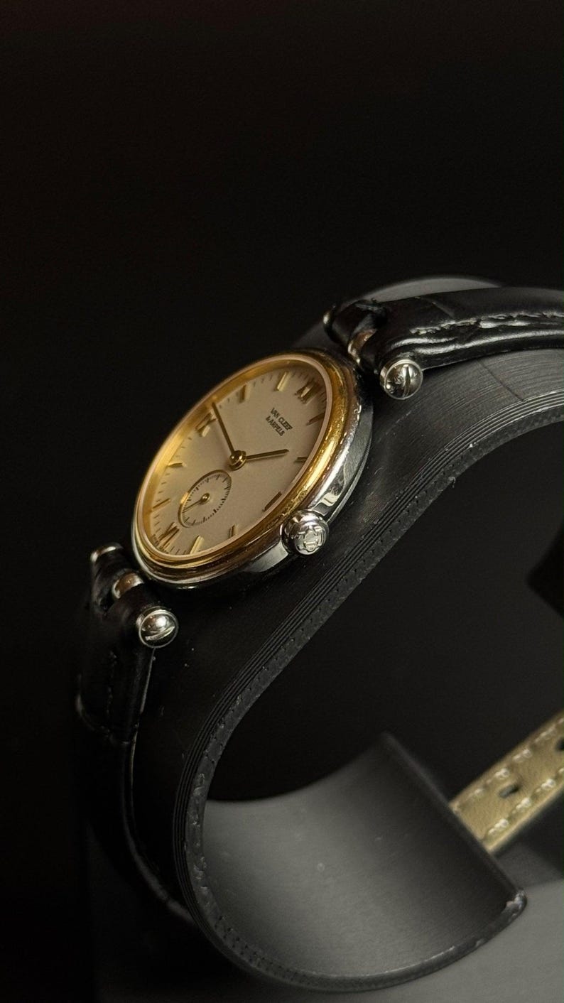 May include: A wristwatch featuring a round silver-tone case with a gold-tone bezel, a cream-colored dial with gold-tone hands and indices, and the text Van Cleef & Arpels. The watch includes a black leather strap with silver-tone hardware.