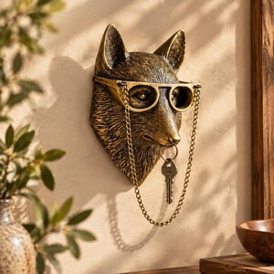 Puede incluir: Una escultura de pared con forma de cabeza de zorro con acabado en bronce metálico. El zorro lleva unas gafas integradas con una cadena metálica fina y larga que cuelga de la montura. Una llave metálica cuelga de una anilla en su boca.