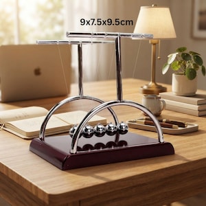 May include: A Newton's cradle featuring five polished silver-colored metal spheres suspended by thin wires from a chrome-finished arched frame. The device is mounted on a dark mahogany-colored wooden base and measures 9x7.5x9.5cm.