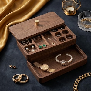 Op de afbeelding: Een rechthoekige donkerbruine houten juwelendoos met een afneembaar deksel voorzien van een kleine gouden knop. Het interieur bevat meerdere vakken, waaronder ronde ringhouders, rechthoekige secties voor kettingen en een uitschuifbare lade.