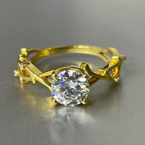 Peut inclure: Une bague en or ornée d'une pierre précieuse ronde, transparente et à facettes au centre. L'anneau présente un motif texturé rappelant des branches qui entoure le doigt, intégrant de petites pierres d'accent incrustées sur la structure métallique.