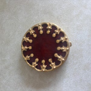 May include: A circular brooch featuring a deep red centre surrounded by a gold-toned metal border. The border is decorated with a repeating pattern of small, ornate, fleur-de-lis style metal accents that frame the dark red circular inlay.