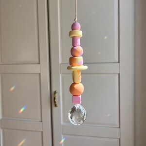 May include: A hanging ornament featuring a vertical string of assorted beads in pastel pink, yellow, and orange hues, including solid, marbled, and geometric shapes, finished with a clear, multifaceted crystal sphere at the bottom end.