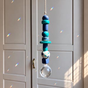 May include: A hanging ornament featuring a vertical arrangement of various beads in shades of navy blue, teal, and white with speckled patterns, suspended by a light-colored cord and finished with a clear, multifaceted crystal sphere at the bottom.