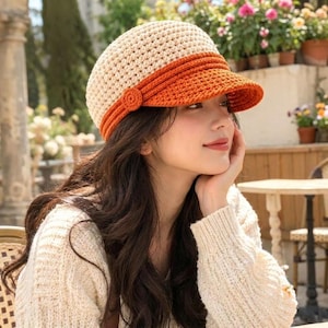 May include: A two-toned crocheted hat featuring a cream-colored crown and a vibrant orange brim. The hat is constructed with a dense, textured stitch pattern and includes a small, circular crocheted button detail on the side of the brim.