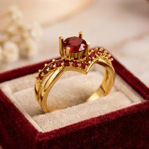May include: A gold ring featuring a prominent round-cut red gemstone at the centre, flanked by a V-shaped band adorned with a row of smaller red stones. The ring has a polished metallic finish and is presented in a deep red velvet jewellery box.