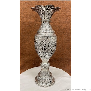 May include: A tall, silver-coloured metal vase featuring an intricate, embossed floral and leaf pattern covering its entire surface. The vase has a wide, flared, petal-shaped rim, a bulbous body, and a circular pedestal base. Text reads: ANCIENT SPIRIT OBJECTS.