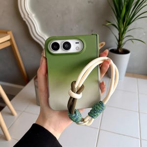 May include: A foldable smartphone case featuring a green-to-light-gray gradient finish. The case includes a white cord wrist strap adorned with a dark olive green knot and two textured, multi-colored blue and green decorative cord wraps.