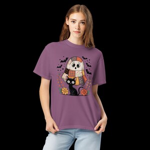 Könnte beinhalten: Ein malvenfarbenes Kurzarm-T-Shirt mit einem Grafikdruck eines Patchwork-Geistes und einer schwarzen Katze. Das Design enthält kleine Fledermäuse, florale Elemente und herbstfarbene Muster auf dem Geist vor einem einfarbigen Stoffhintergrund.
