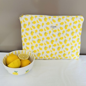 Peut inclure: Une pochette matelassée rectangulaire avec un motif répétitif de citrons jaunes sur fond blanc, une fermeture éclair dorée et une étiquette en tissu marquée Frenchic Création, accompagnée d'un bol blanc contenant trois citrons entiers et coupés.