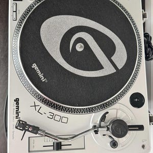 May include: A silver Gemini XL-300 fully manual direct-drive turntable featuring a black slipmat with a large grey logo. The device includes a chrome tonearm, a pitch adjustment slider, speed selection buttons, and a power switch.