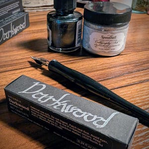 May include: A black calligraphy pen, a small glass bottle with a dropper, and a jar of Speedball silver calligraphy ink. A rectangular black box labeled Darkwood with 0.34 fl oz text is positioned in the foreground on a wooden surface.