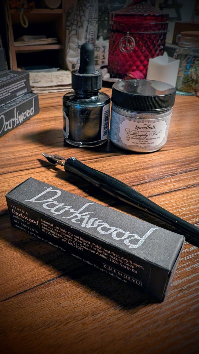 May include: A black calligraphy pen, a small glass bottle with a dropper, and a jar of Speedball silver calligraphy ink. A rectangular black box labeled Darkwood with 0.34 fl oz text is positioned in the foreground on a wooden surface.