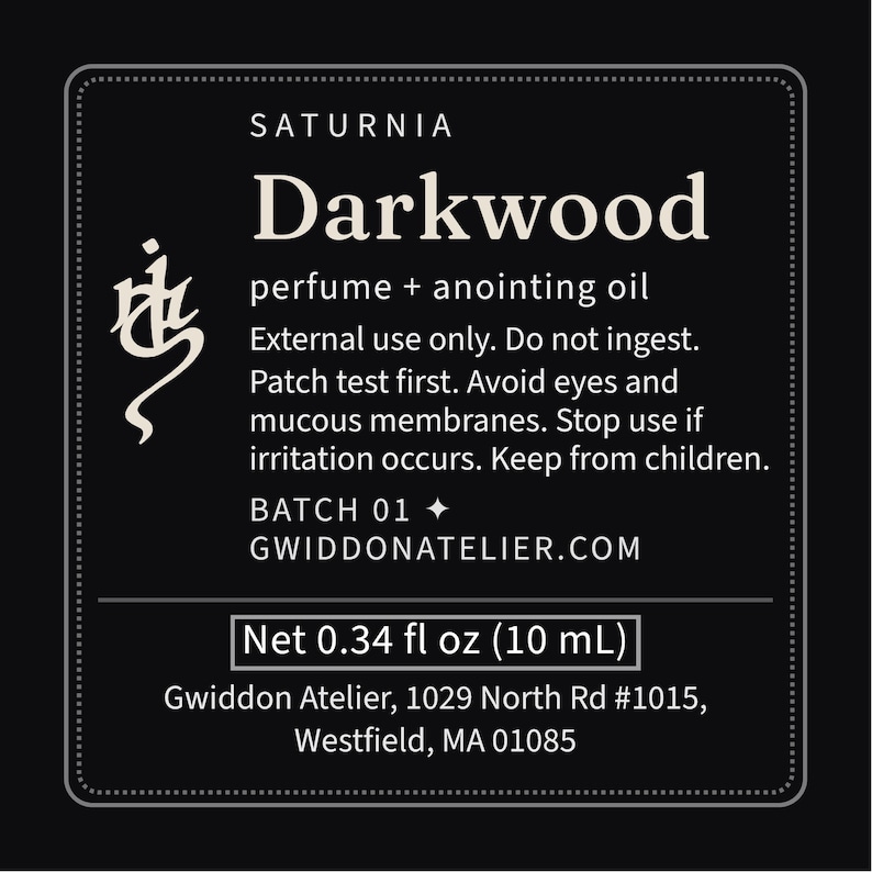 May include: A black rectangular product label with white text reading Saturnia Darkwood perfume + anointing oil. It includes safety warnings, Batch 01, the website gwiddonatelier.com, and the net volume of 0.34 fl oz (10 mL) with address details.
