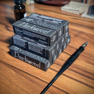 May include: A stack of three black rectangular boxes labeled Darkwood with white text and a black dip pen with a metal nib. The boxes feature the text Saturnia and list ingredients including jojoba oil, ginger, and various essential oils in 0.34 fl oz.