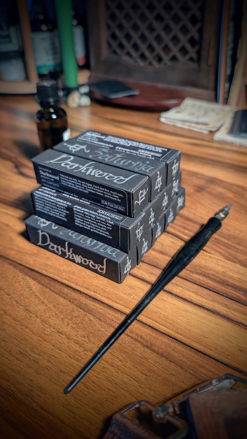 May include: A stack of three black rectangular boxes labeled Darkwood with white text and a black dip pen with a metal nib. The boxes feature the text Saturnia and list ingredients including jojoba oil, ginger, and various essential oils in 0.34 fl oz.
