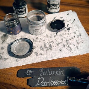 May include: A collection of calligraphy supplies including a bottle of Dr. Ph. Martin's Iridescent ink, a jar of Speedball silver ink, and a bottle of India ink. Two black paper strips feature the handwritten words Saturnia and Darkwood in silver.