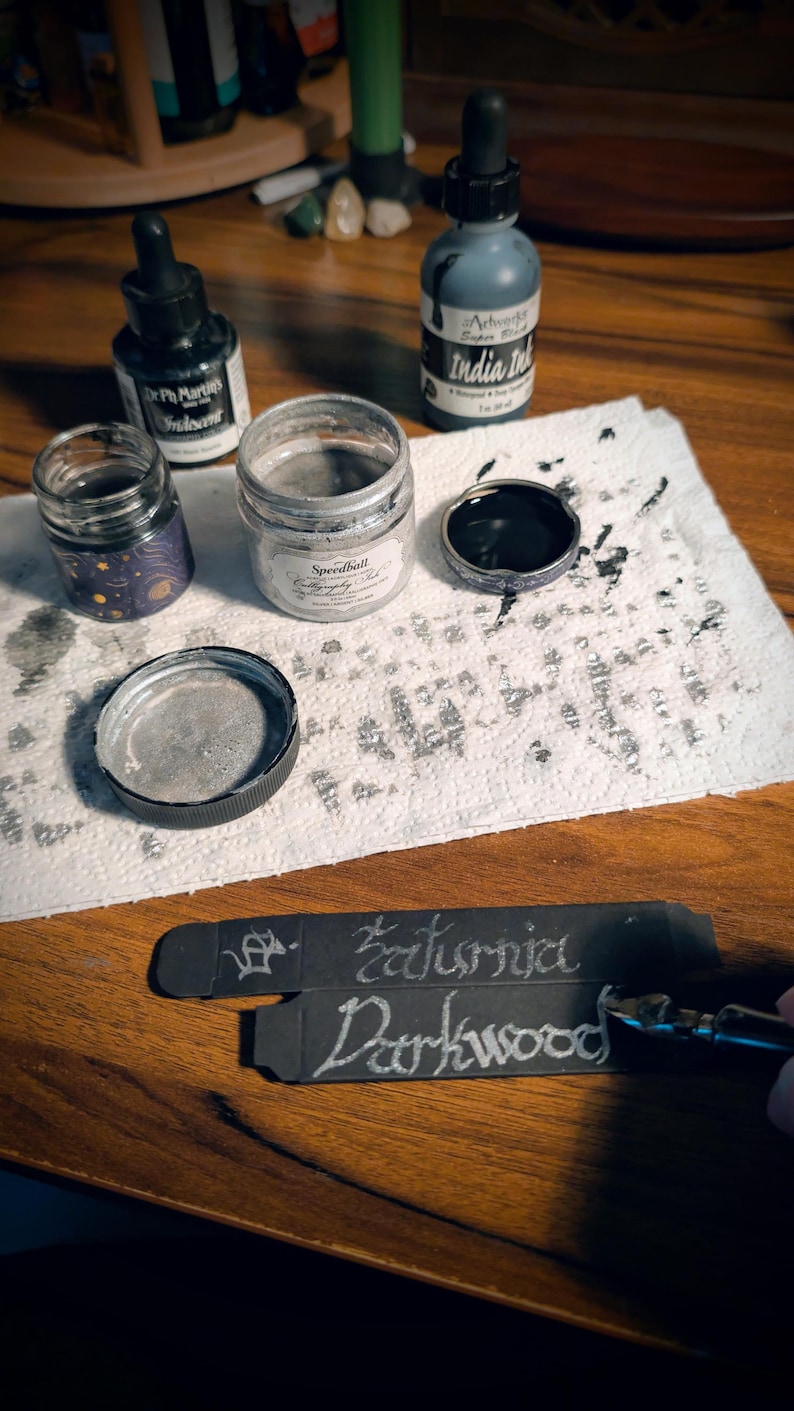 May include: A collection of calligraphy supplies including a bottle of Dr. Ph. Martin's Iridescent ink, a jar of Speedball silver ink, and a bottle of India ink. Two black paper strips feature the handwritten words Saturnia and Darkwood in silver.