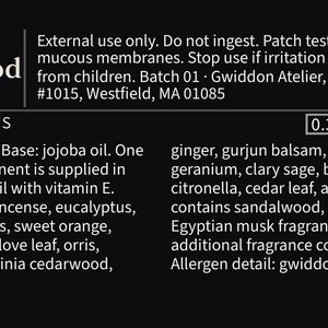 May include: A black product label for Saturnia Darkwood perfume and oil, 0.34 fl oz. Text includes safety warnings, batch 01, Gwiddon Atelier address, and a list of ingredients such as jojoba oil, frankincense, eucalyptus, patchouli, and sandalwood.