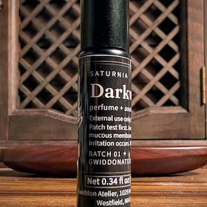 May include: A black cylindrical perfume roller bottle with a label reading SATURNIA Dark, perfume + anointing oil, External use only, Patch test first, Avoid mucous membranes, if irritation occurs, BATCH 01, GWIDDONATEL, Net 0.34 fl oz, Gwiddon Atelier.