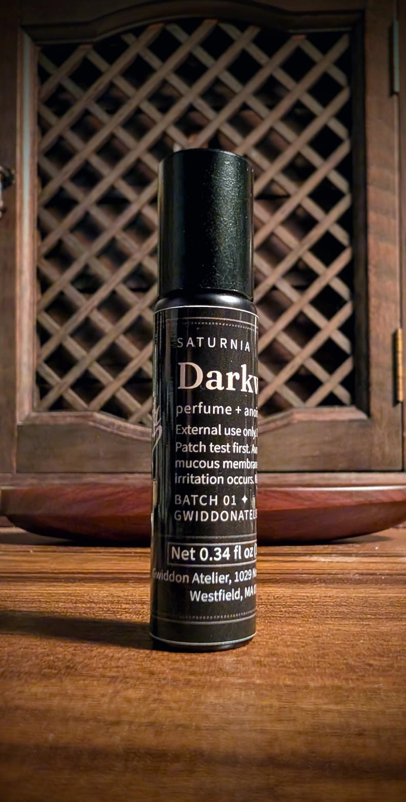 May include: A black cylindrical perfume roller bottle with a label reading SATURNIA Dark, perfume + anointing oil, External use only, Patch test first, Avoid mucous membranes, if irritation occurs, BATCH 01, GWIDDONATEL, Net 0.34 fl oz, Gwiddon Atelier.