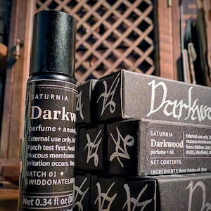 May include: A black cylindrical perfume bottle with a label reading Saturnia Darkwood perfume + anointing oil, Net 0.34 fl oz, alongside stacked black rectangular boxes featuring the text Darkwood and white hand-drawn sigil-like symbols.