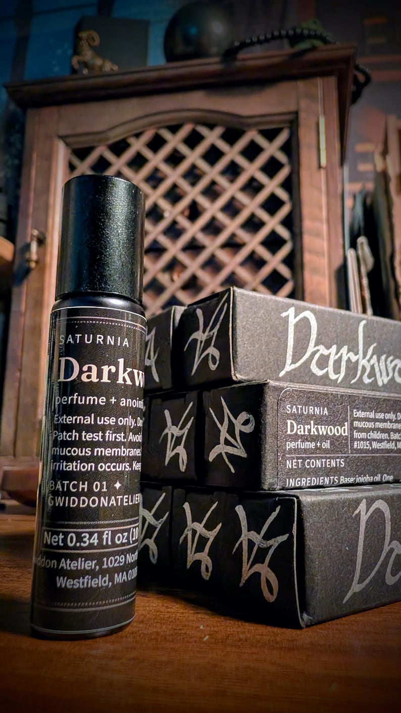 May include: A black cylindrical perfume bottle with a label reading Saturnia Darkwood perfume + anointing oil, Net 0.34 fl oz, alongside stacked black rectangular boxes featuring the text Darkwood and white hand-drawn sigil-like symbols.