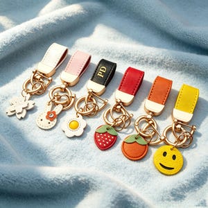 Op de afbeelding: Zes sleutelhangers met goudkleurige metalen hardware en kunstlederen bandjes in wit, roze, zwart, rood, oranje en geel. De bedels bestaan uit een witte bloem, een konijn, een wit madeliefje, een aardbei, een sinaasappel en een geel smiley-gezicht.