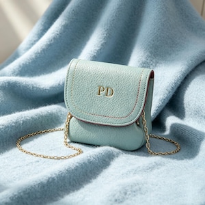May include: A small, light blue textured crossbody bag with a flap closure featuring the gold embossed letters PD. The bag has a thin gold metal chain strap and visible red stitching along the edge of the flap, creating a distinct contrast.