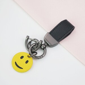 Op de afbeelding: Een sleutelhanger met een felgele, ronde smiley-bedel bevestigd aan een donkergrijze metalen hardware-constructie, inclusief een stevige ring en een zwarte, gestructureerde leren lus voor bevestiging aan sleutels of tassen.