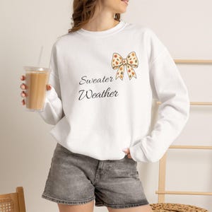 Könnte beinhalten: Ein weißes langärmeliges Sweatshirt mit Rundhalsausschnitt, das eine Grafik einer Schleife mit Herbstblattmuster und den darunter gedruckten schwarzen kursiven Text Sweater Weather zeigt. Das Sweatshirt wird mit grauen Jeansshorts kombiniert.